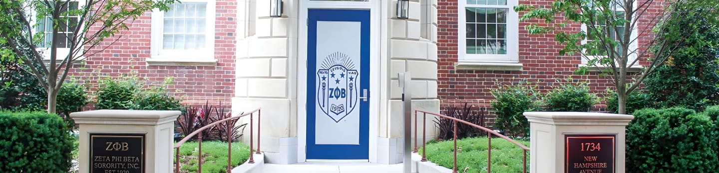 Stone pillars with plaques flank a brick building with a blue-trimmed door and windows, surrounded by manicured bushes and trees.