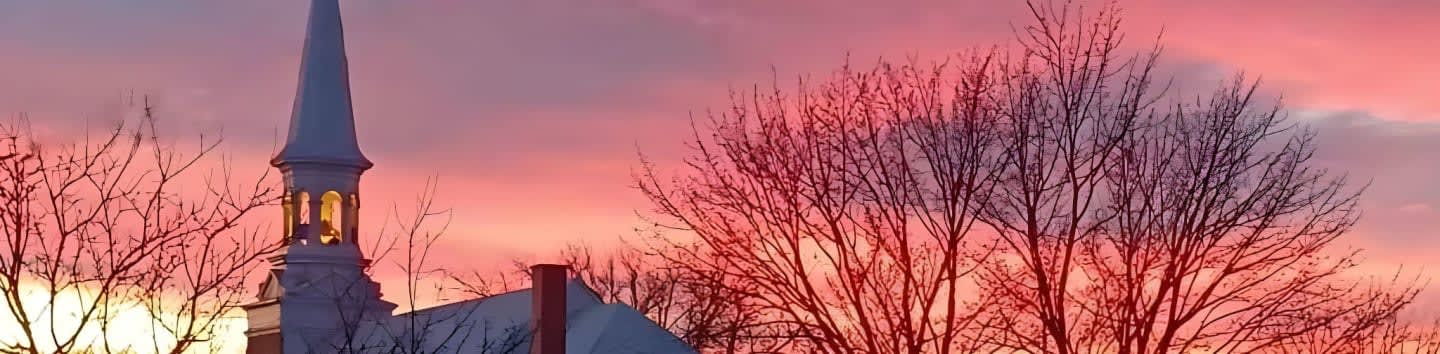 Bare trees frame a white steeple against a vibrant pink and orange sunset sky.