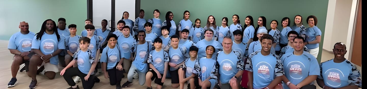 A large group of people, mostly children and young adults, are posing for a photo in light blue t-shirts in front of a light green wall.