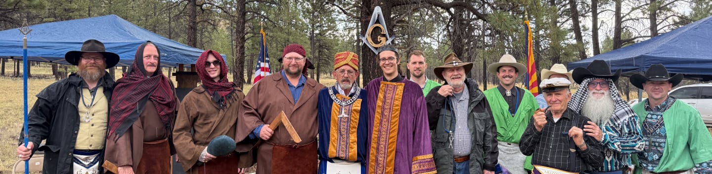 A group of men in various historical and ceremonial attire stand in a wooded area with tents and trees in the background.