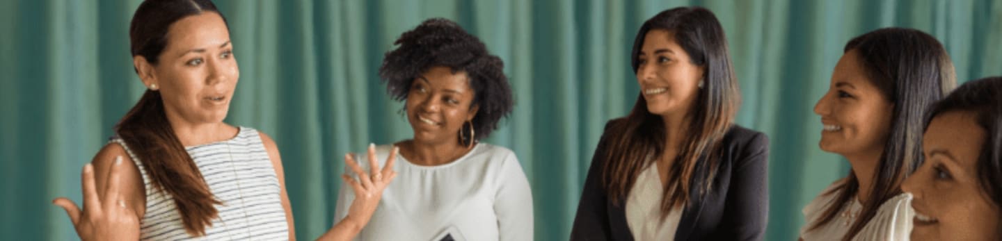 A group of diverse women engage in conversation at a networking meetup, with a teal curtain in the background.