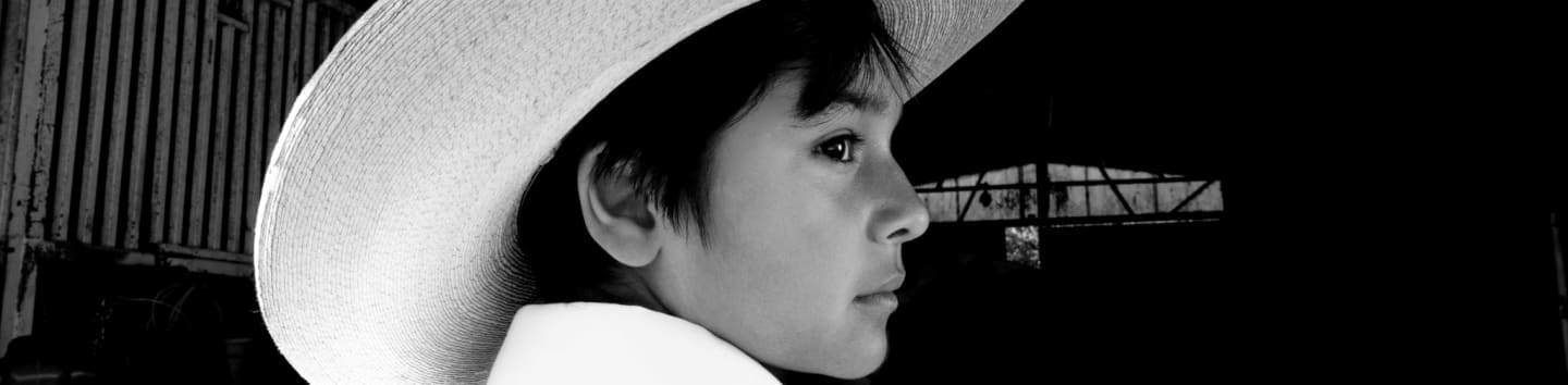 A young boy in a cowboy hat looks out from a rustic building towards a dark, industrial background.