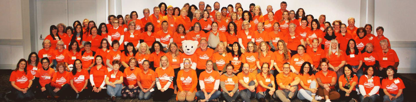 A large group of people in orange shirts are posing for a photo in front of a plain background, with a mascot in the center.