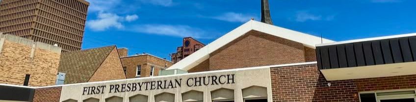 The "First Presbyterian Church" sign is visible on the front of the brick building, with a tall, brown, grid-like skyscraper in the background under a bright blue sky.