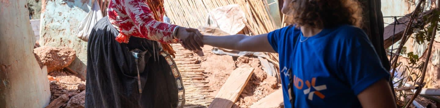 Two people shake hands in the foreground, with a backdrop of rubble and debris.
