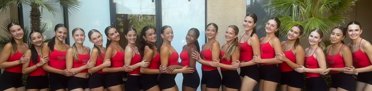 A group of young dancers in red tops and black shorts stand in front of a building with palm trees.