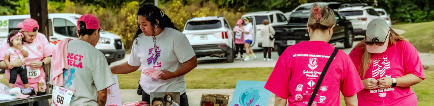 People in pink shirts are gathered around a table in the foreground, with cars parked in the background.