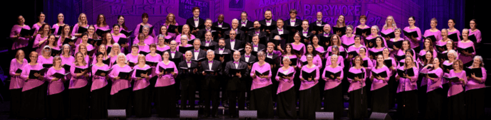A large choir in pink tops and black bottoms stands on a stage with a purple, blurred background.