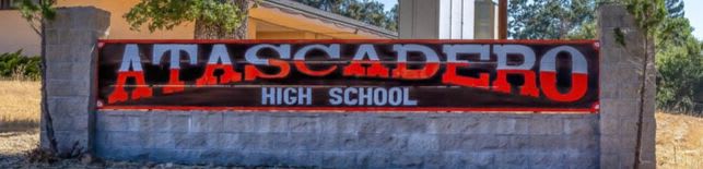 A weathered stone monument with the "ATASCADERO HIGH SCHOOL" sign stands before a building and trees under a clear sky.
