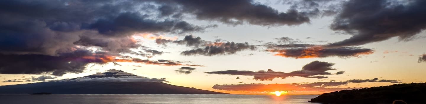 A calm ocean reflects a dramatic sunset with dark clouds and a distant island.