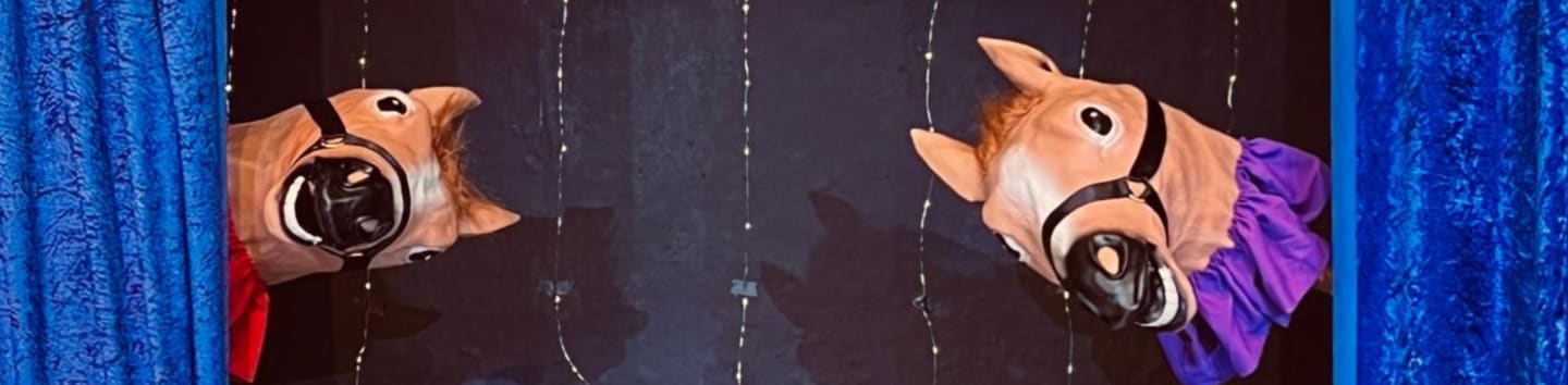 Two horse puppets with brown heads and black harnesses peek out from behind blue curtains, with faint lights strung in the dark background.