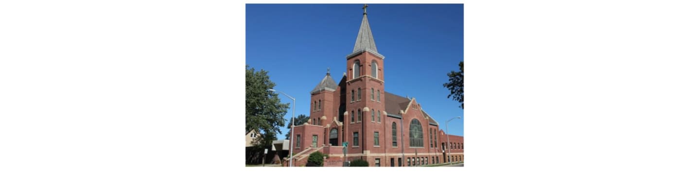 A brick church with a tall steeple stands against a clear blue sky, with trees visible on either side.