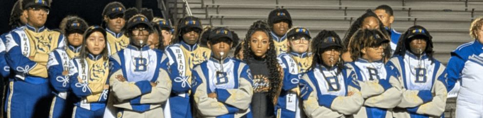 A marching band in blue and gold uniforms stands on a grassy field in front of bleachers at night.