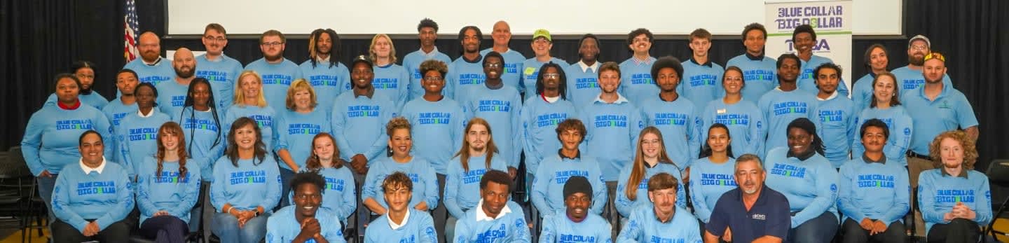 A group of people in light blue sweatshirts are posing for a photo in front of a black curtain and a white banner.