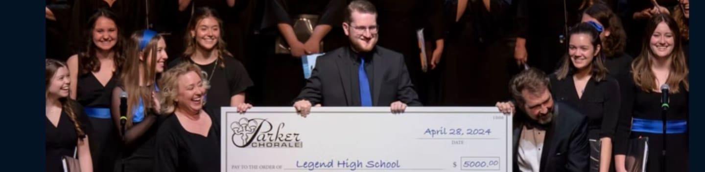 A group of smiling people in formal attire hold a large check in front of a choir.