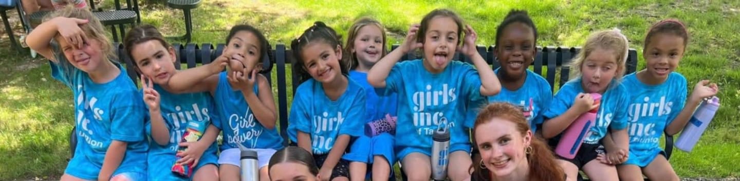 A group of young girls in blue "Girls Inc." t-shirts pose for a photo on a bench and on the ground in front of a grassy, tree-lined background.