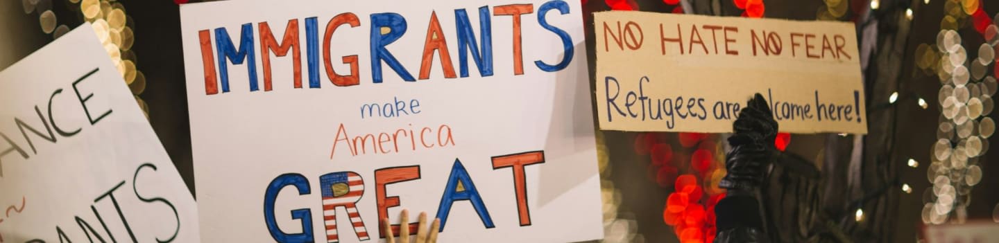 Handwritten signs advocating for immigrants and refugees are displayed against a blurred background of lights.