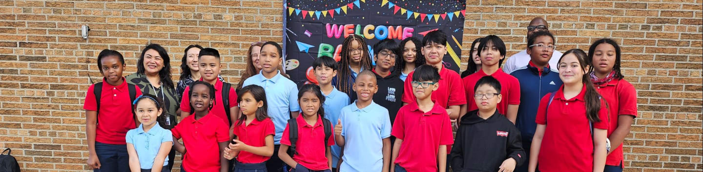 A group of students and adults pose in front of a brick wall with a "Welcome" banner.