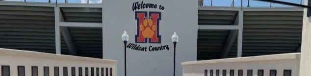 A beige building with a row of windows is in the foreground, while a stadium with a large "H" logo and the words "Welcome to Wildcat Country" is in the background.