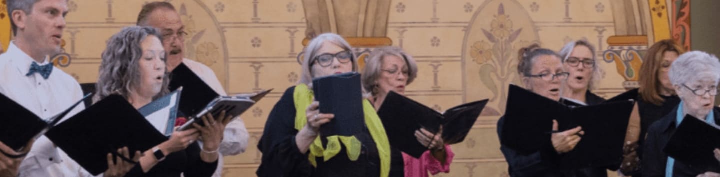 A choir of people in black attire sings from sheet music in front of a large, ornate mural.