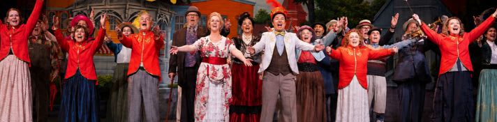 A group of performers in colorful costumes with their arms raised are on a stage with a painted backdrop of buildings.