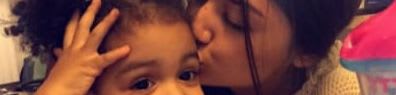 A young girl with curly hair smiles as an older girl kisses her cheek, with a blurry background of a room.