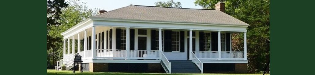 A white house with a wraparound porch sits on a grassy lawn, surrounded by lush green trees.