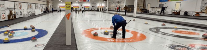 In the foreground, yellow curling stones rest on a yellow mat, while in the background, a person in a blue shirt sweeps the ice on a curling rink with other stones scattered across it.