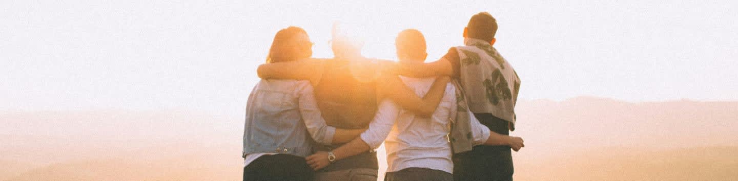 Four friends embrace each other with arms around shoulders against a bright, hazy sky.