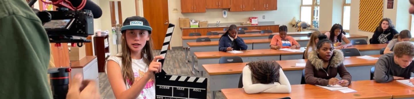 A young girl holds a clapperboard in front of a classroom of students working at desks.