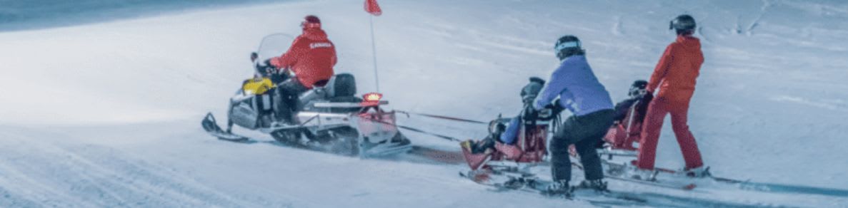 A snowmobile tows a sled with people on skis down a snowy slope at night, with ski lifts and lights illuminating the background.