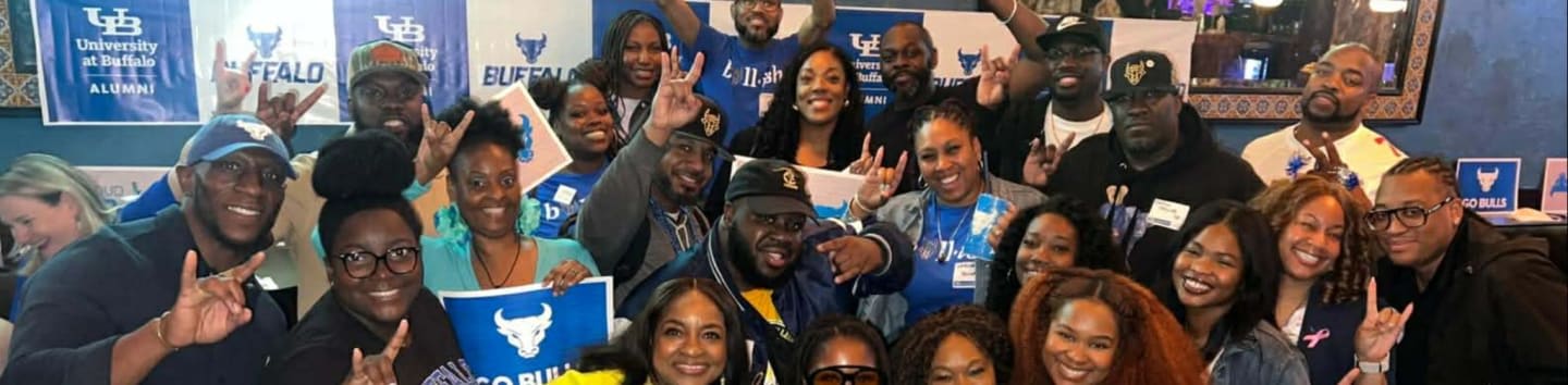 A diverse group of smiling people, many wearing blue and white, pose for a photo in front of banners with the University at Buffalo logo.