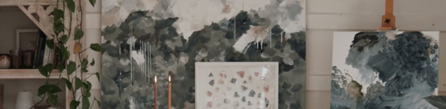 Several women are gathered around a table in a bright art studio, working on their paintings with large abstract art pieces hanging on the wall behind them.