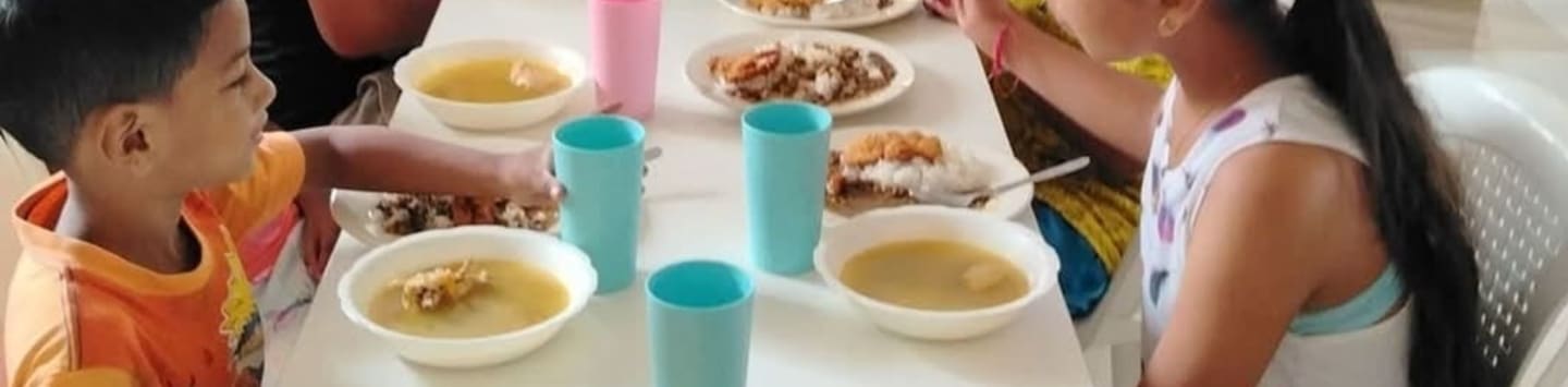 Two children eat soup and rice at a table with blue cups and pink cups in the background.
