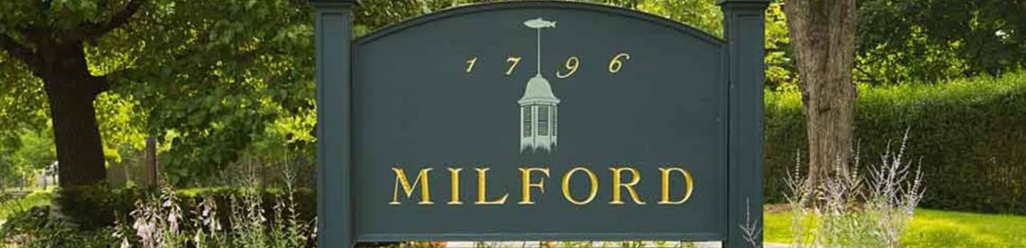 A dark green sign with "MILFORD" and "1796" in gold lettering stands in front of lush green trees and manicured hedges.