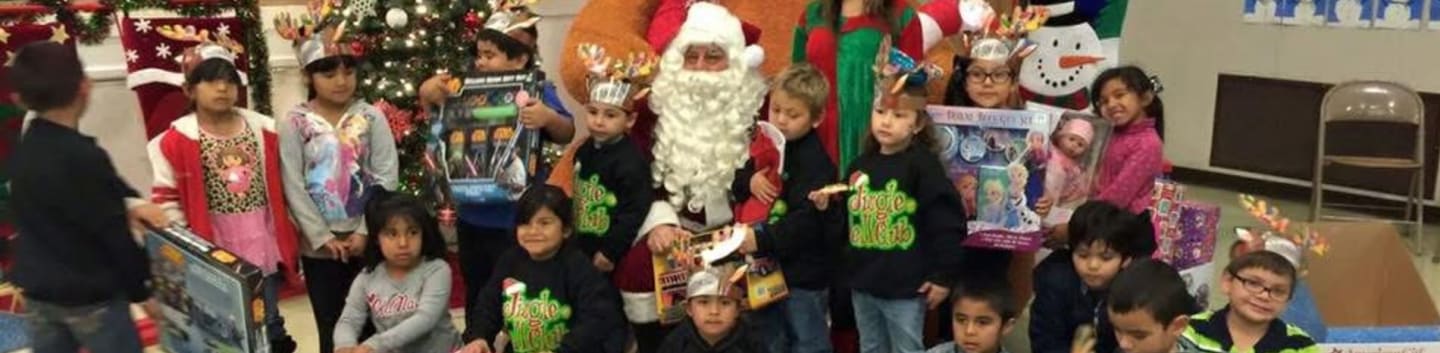 Children in festive attire gather around Santa and an elf, with a decorated Christmas tree and a snowman in the background.