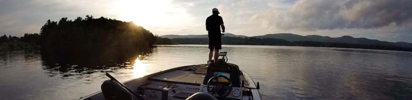 A lone figure stands on the bow of a boat in the foreground, facing a calm lake and distant, tree-covered hills under a cloudy sky.