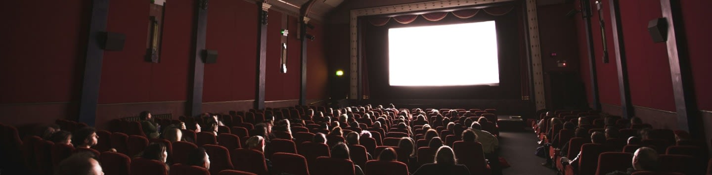 An audience in a dark cinema with red velvet seats facing a bright screen