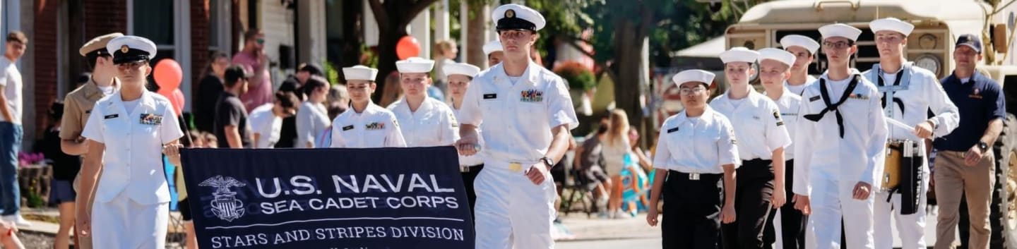 A group of U.S. Naval Sea Cadets march in a parade with a banner, while spectators watch from the background.