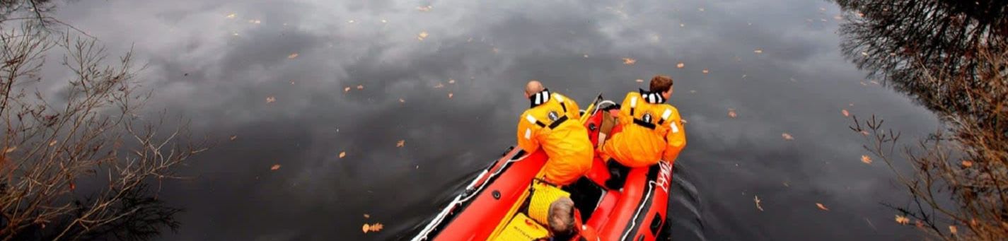 Two people in orange life vests are in a red inflatable boat on a dark, reflective body of water with bare trees and fallen leaves on the banks.