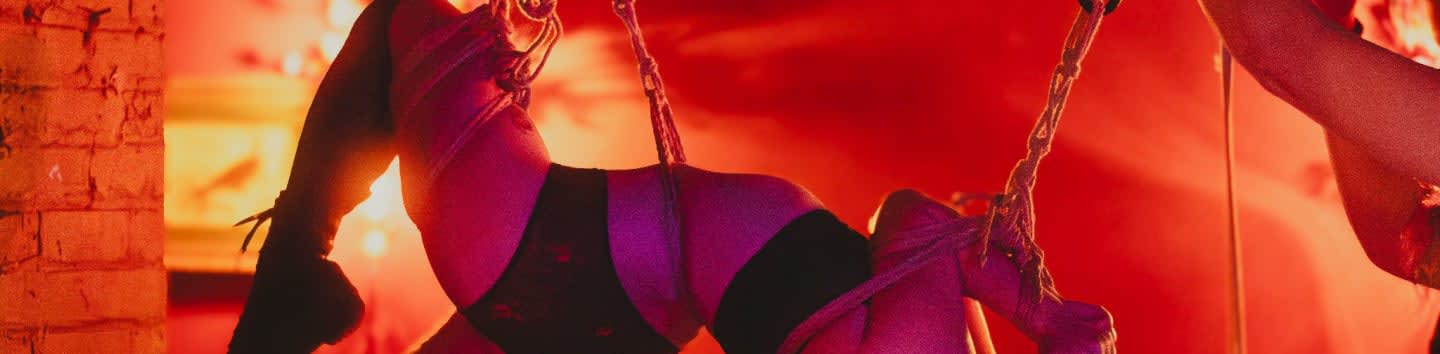 A person in black lingerie is bound by ropes in the foreground against a glowing red and orange background.