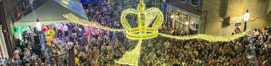A large, glowing golden crown decoration hangs over a dense crowd of people gathered in a city street at night.