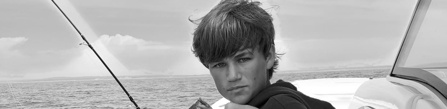 A young boy sits on a boat in the foreground with a fishing rod and a vast ocean under a cloudy sky in the background.
