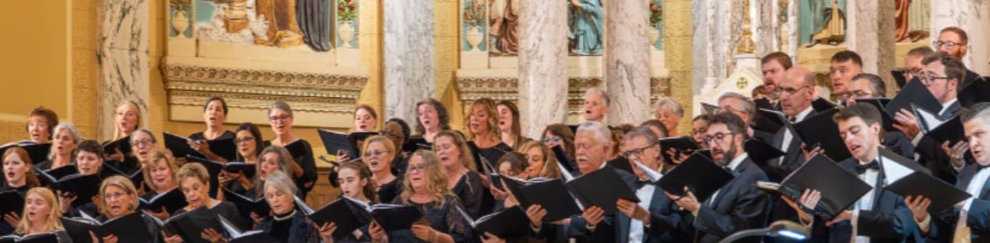 A choir and orchestra perform in a church with ornate arches and religious artwork in the background.