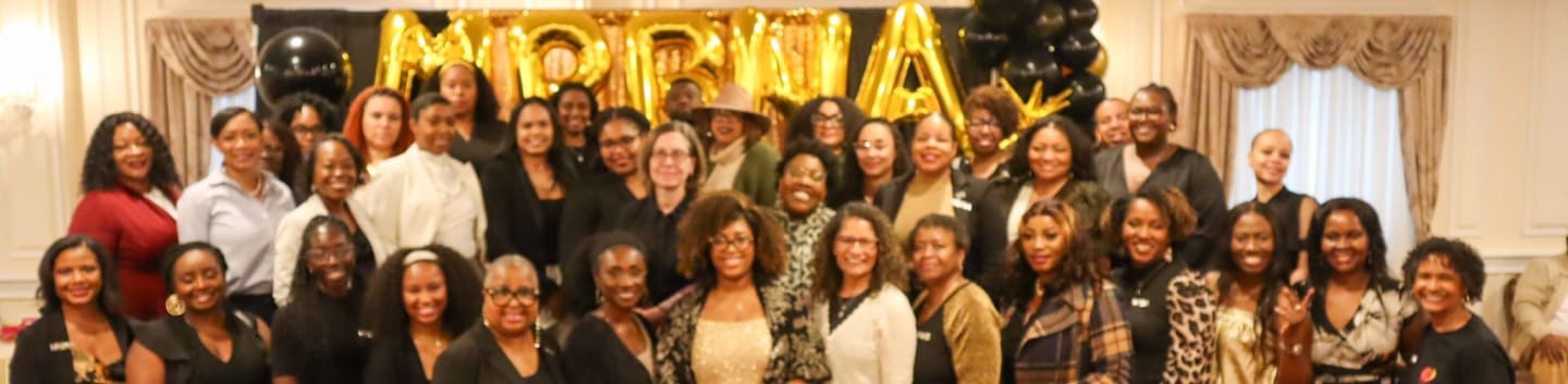 A large group of women pose together in the foreground in front of a black backdrop with gold balloons and lettering in the background.