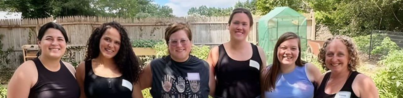 Six women stand in a garden with a wooden fence and greenery in the background.