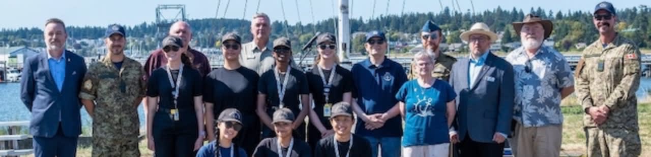 A diverse group of people in military and civilian attire stand together in the foreground against a background of a waterfront town and a bridge.