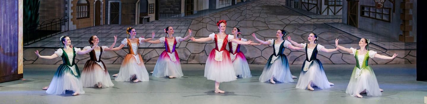 Nine ballet dancers in colorful gradient dresses perform on a stage set with a painted European village backdrop.