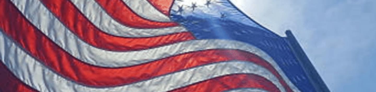 An American flag waves in the foreground against a bright, sunny blue sky.