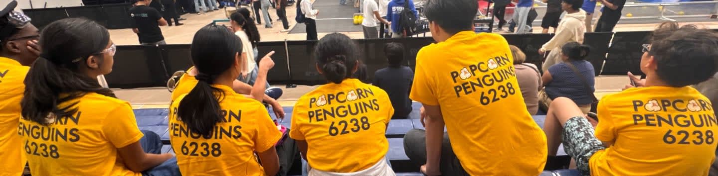 A group of people wearing yellow "Popcorn Penguins 6238" t-shirts sit in the foreground, facing a crowded indoor arena background.